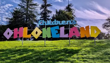 A rainbow-colored sign on a patch of grass under a puffy clouded sky and in front of tall pine trees reads Children's Ohloneland.