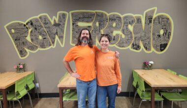 A man and a woman in orange shirts stand smiling in front of a hand-drawn "Raw Fresno" mural inside the restaurant, with wooden tables and green chairs on either side.