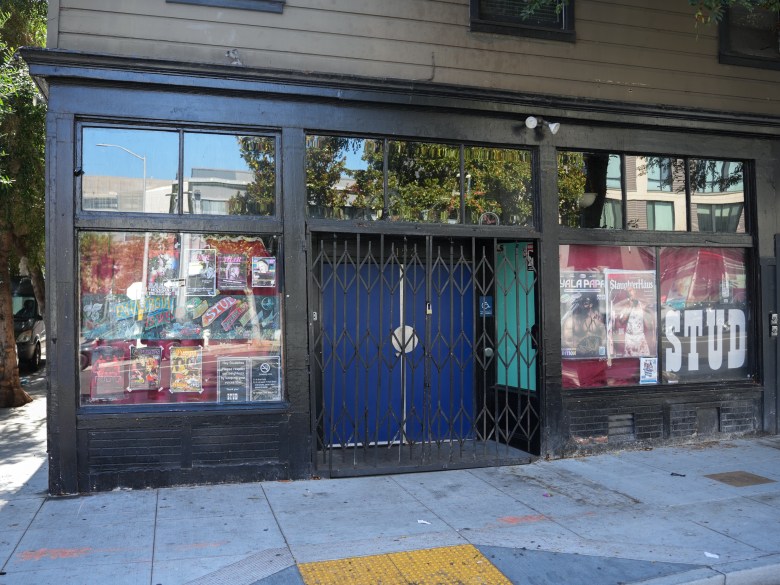 A storefront with large windows, a closed metal security gate over blue doors, and posters on the glass advertising events and the word “STUD.”.