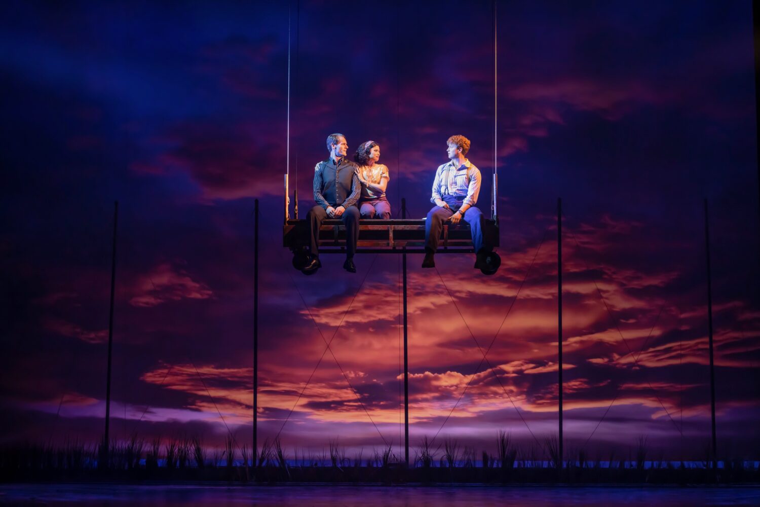 Three adults share a suspended bench ride against a dramatic orange-pink sunset sky, silhouetted poles in the background.