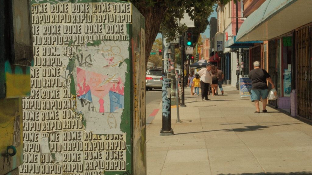 On the right of the frame we see the backs of half a dozen people walking on a sidewalk past storefronts and approaching a greenlight. To the left is a ripped poster on a box with the words Rome and Rapid all over it.
