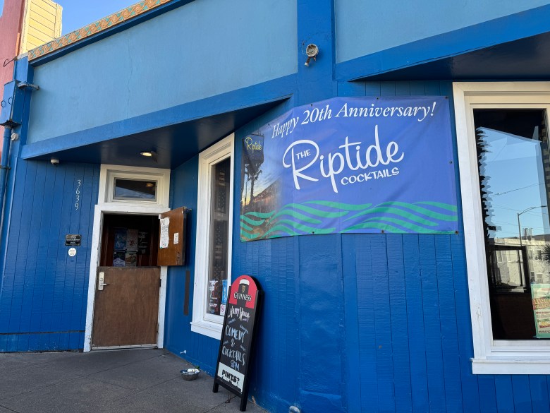 A blue building with a sign reading “Happy 20th Anniversary! The Riptide Cocktails” hangs above the entrance. A smaller chalkboard sign stands on the sidewalk.