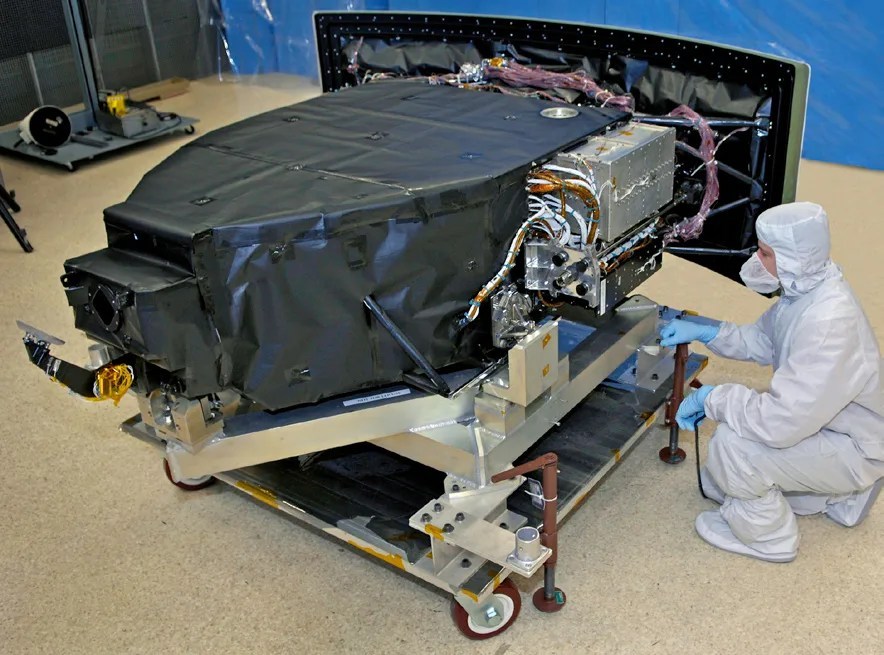 A person in a cleanroom suit kneels beside a large, covered scientific instrument on a wheeled cart, with cables and components exposed, in a laboratory setting. Credit: NASA