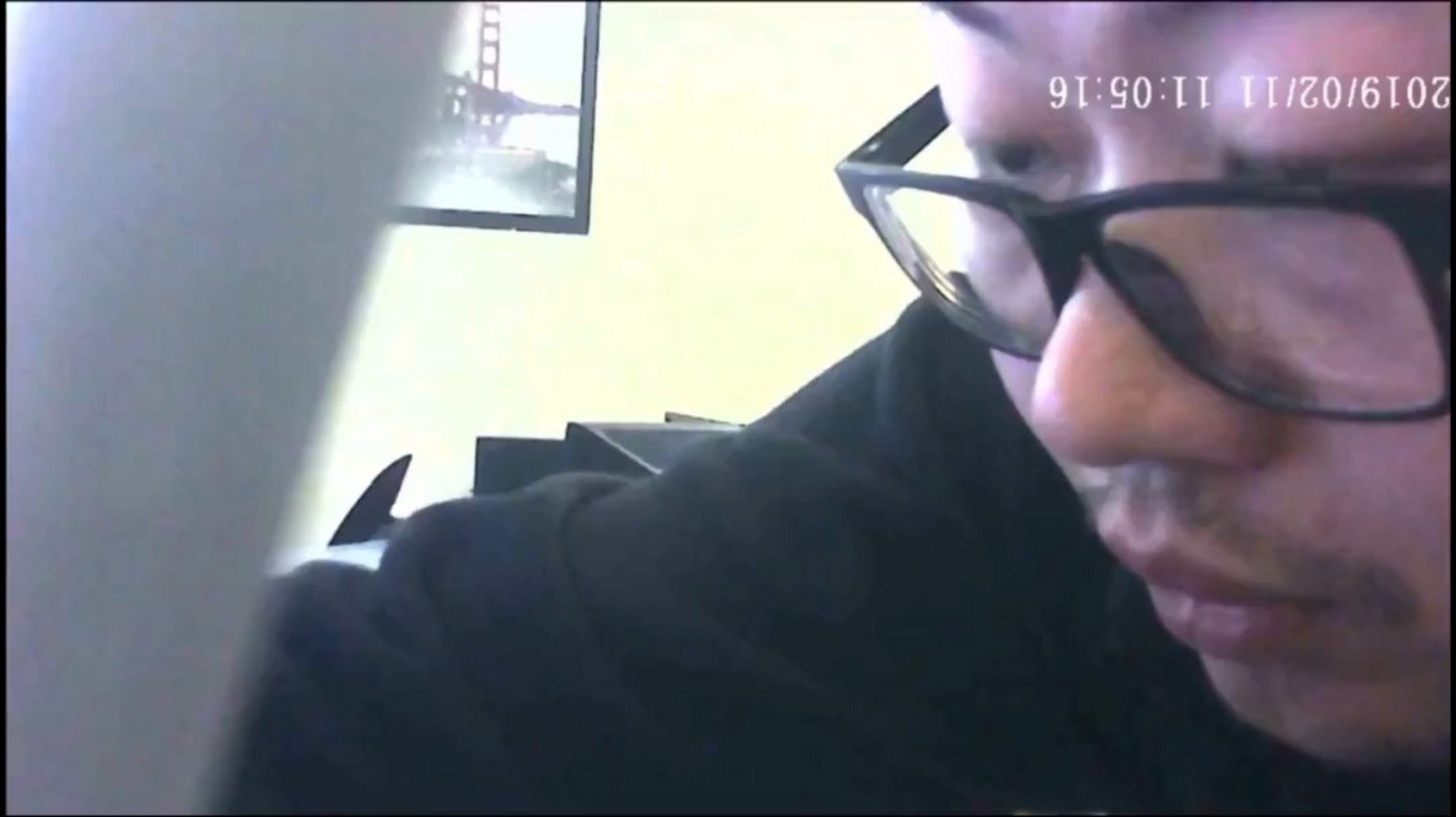 A person wearing black glasses and a black top is leaning forward, partially obscuring the camera, with a picture of the Golden Gate Bridge visible on the wall behind.