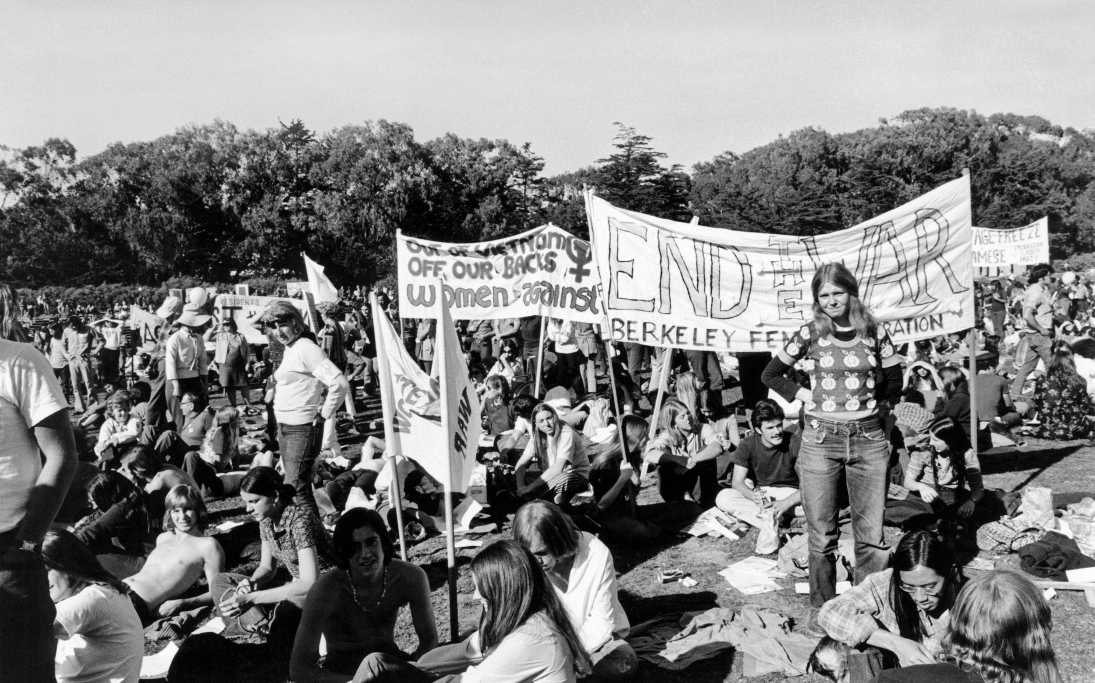 A large group of people sit on grass during a protest, holding signs that include “END THE WAR” and “Women Against War” near trees in the background.