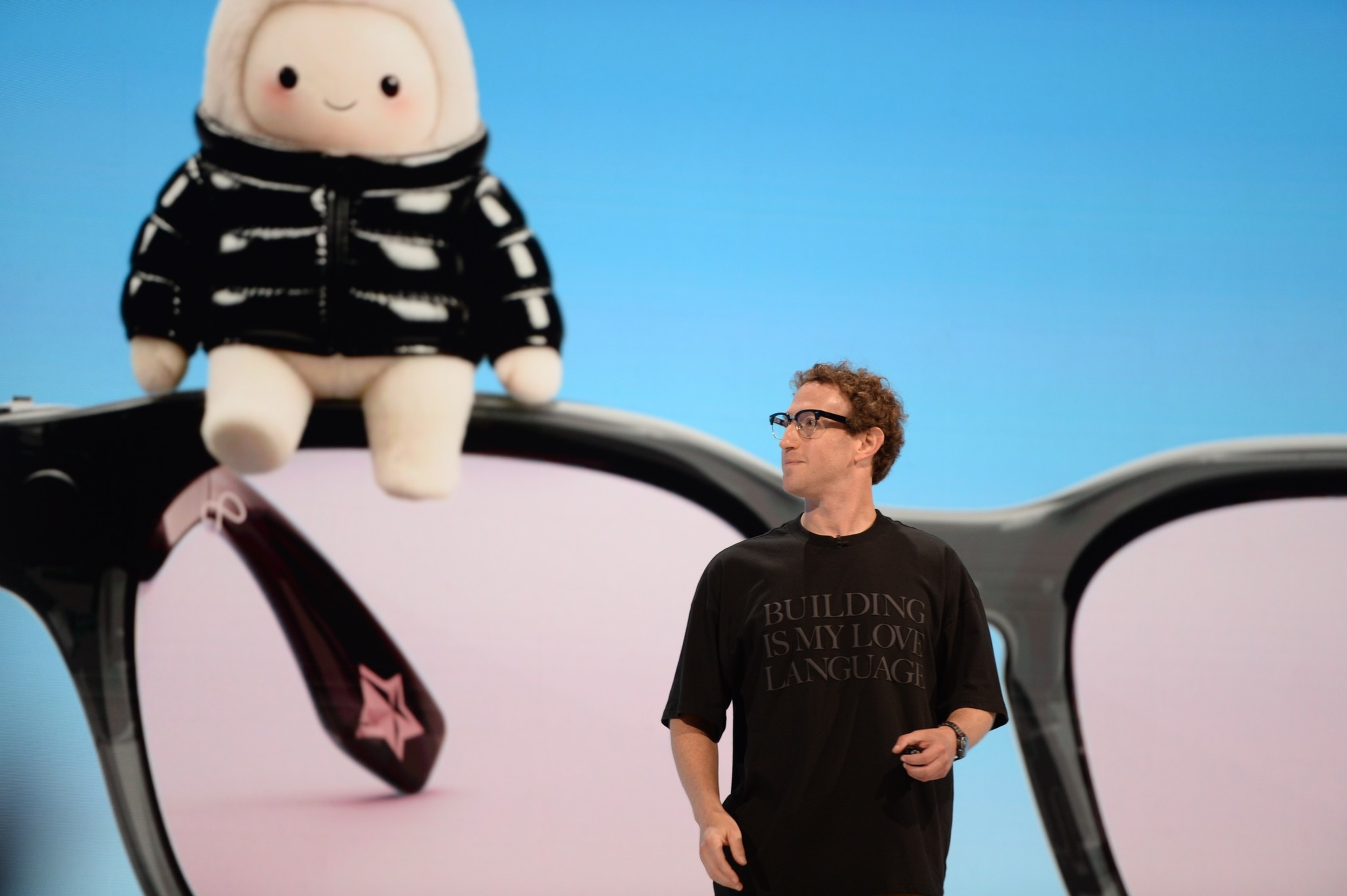A man wearing glasses and a black shirt with “BUILDING IS MY LOVE LANGUAGE” stands near a large image of glasses with a small plush doll in a black jacket.