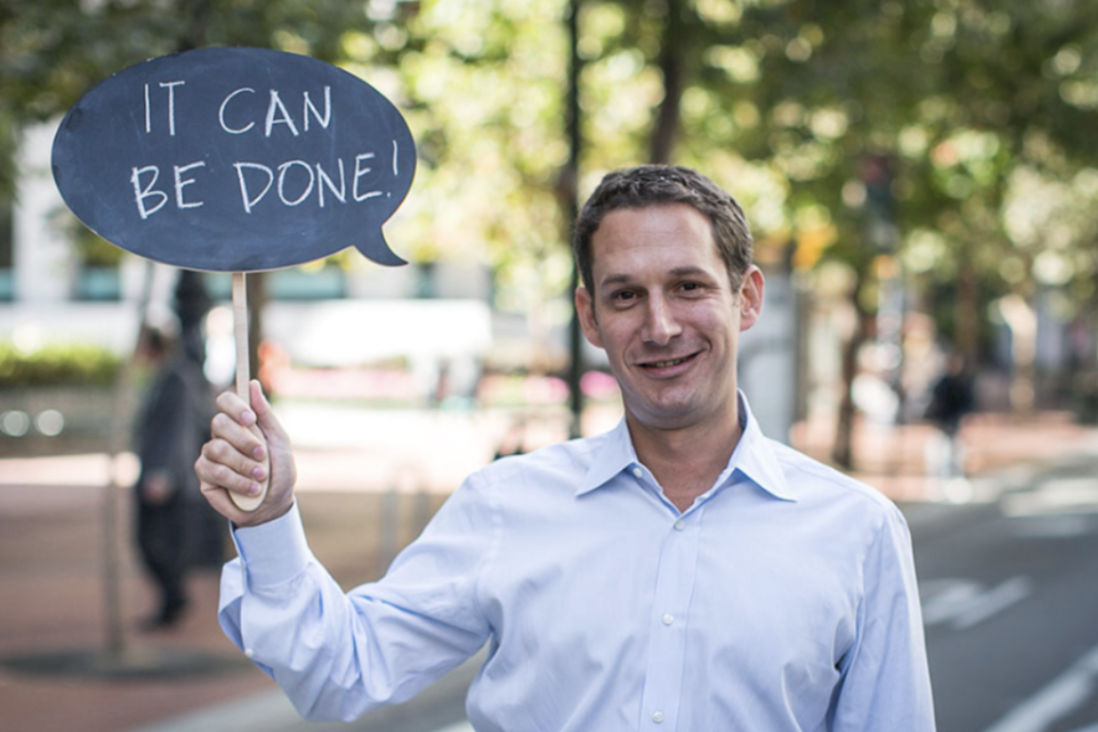 A man in a light blue shirt smiles and holds a speech bubble sign that reads “IT CAN BE DONE!” with a blurred outdoor background.