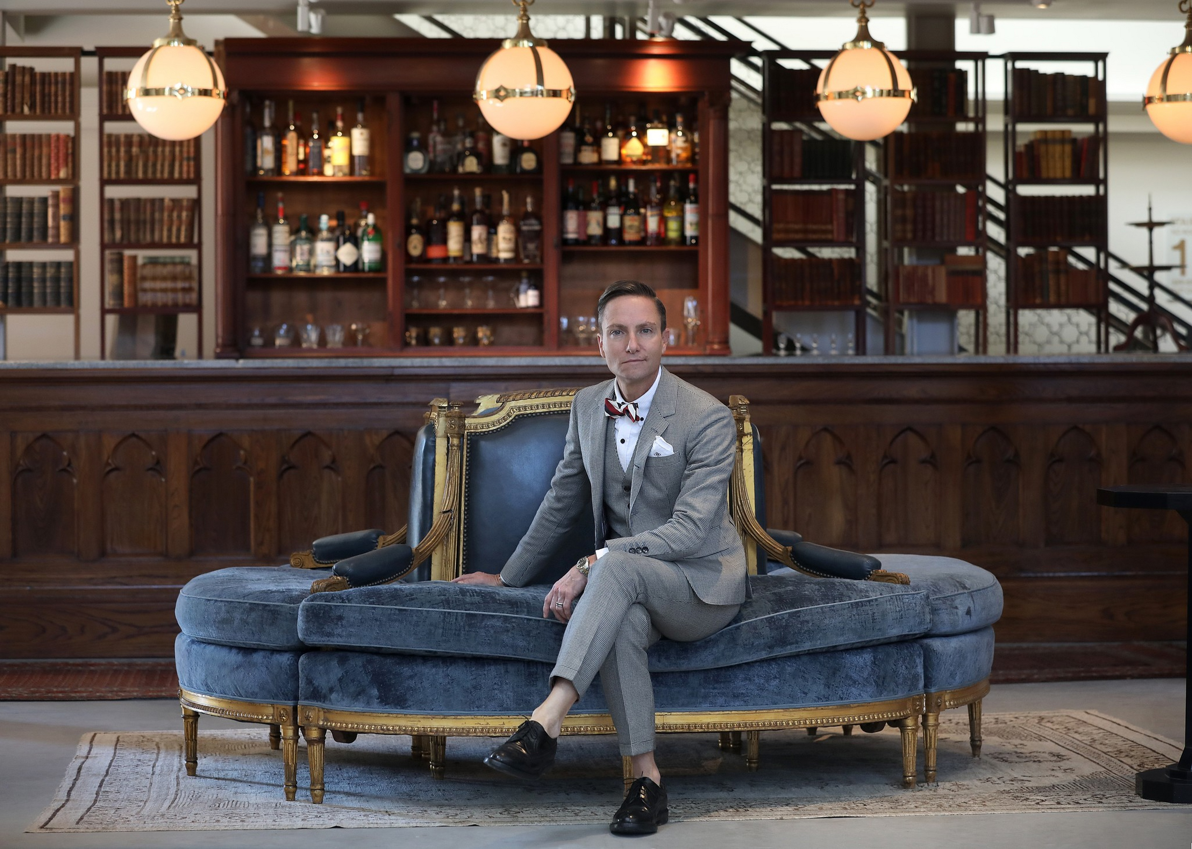 A man in a gray suit and bow tie sits on a luxurious blue velvet circular sofa in front of a wooden bar with shelves of bottles and books.
