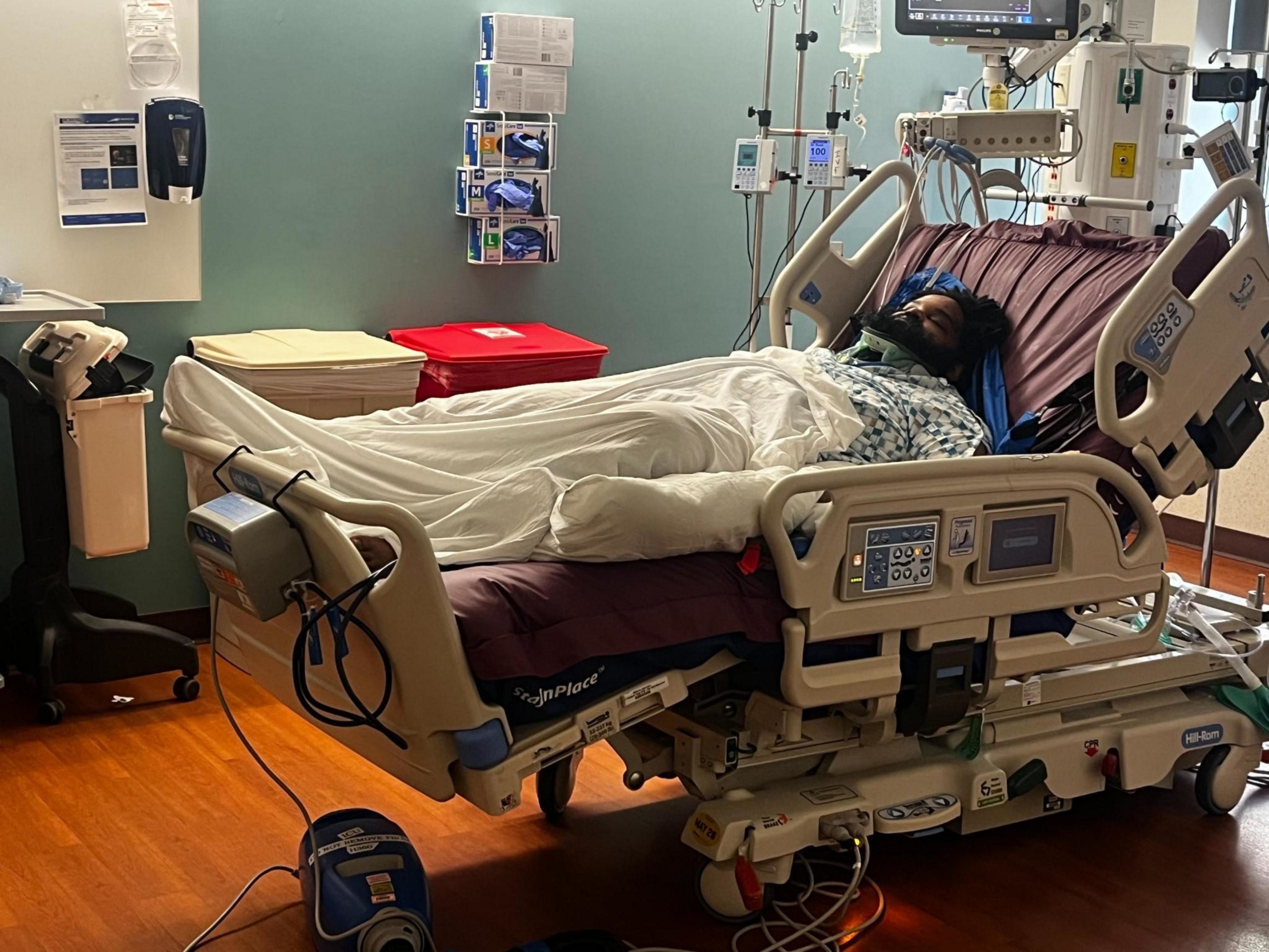 A patient lies in a hospital bed covered with a white blanket, wearing a neck brace and connected to medical monitors and IV drips.