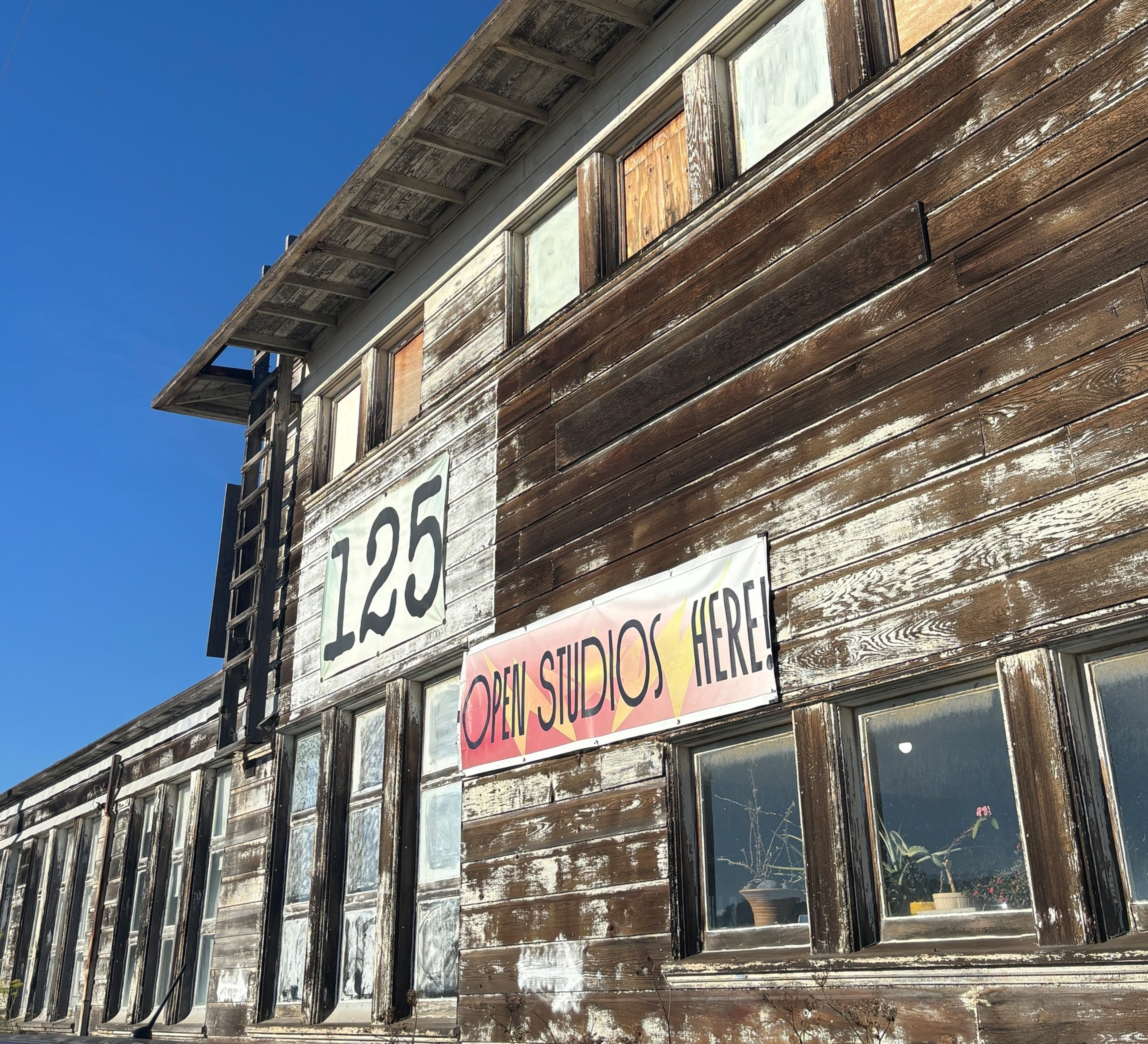 A weathered wooden building with boarded-up upper windows displays the number “125" and a colorful banner reading ”Open Studios Here!” under clear blue sky.