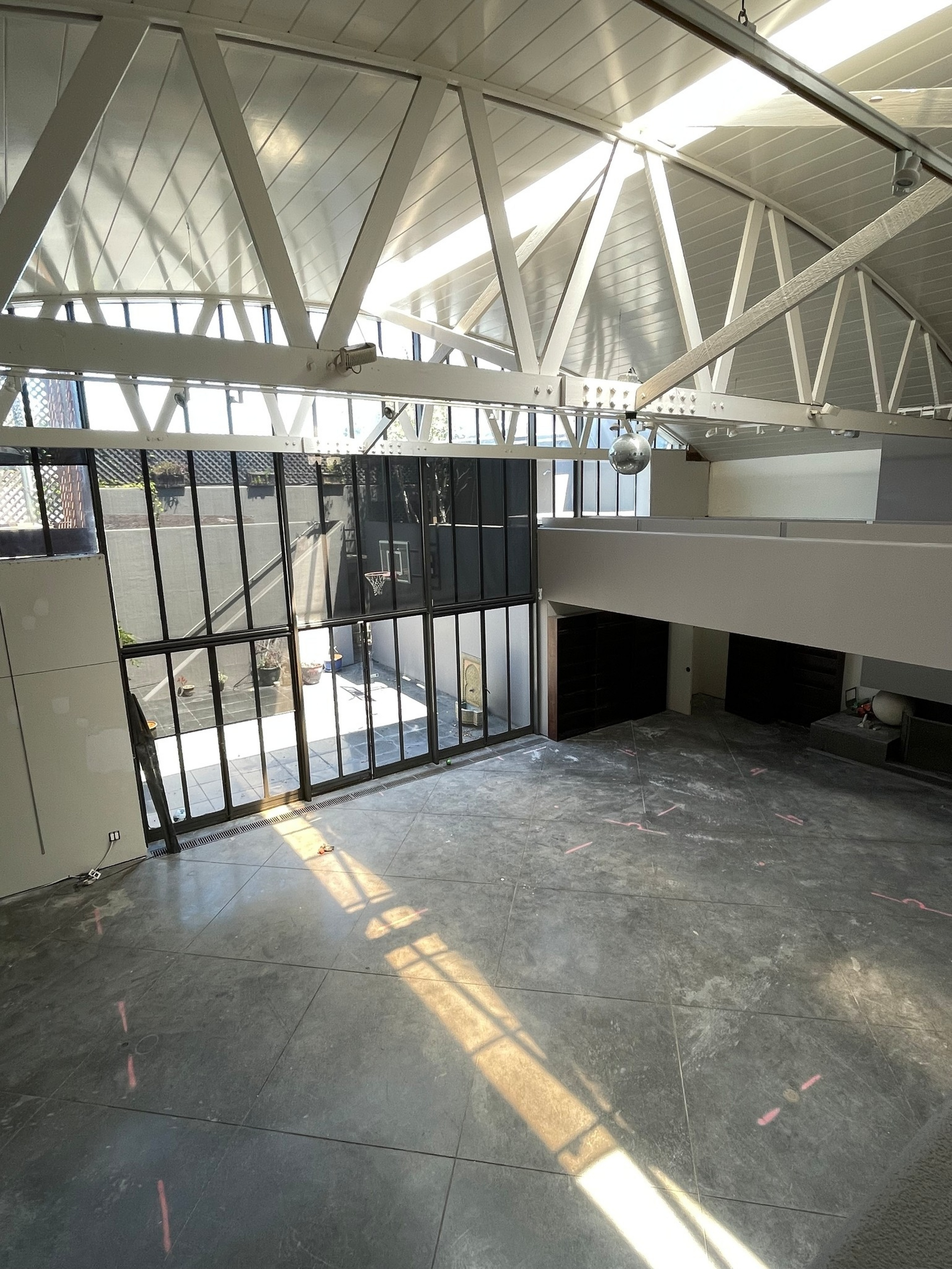 A spacious indoor court with concrete flooring, large industrial windows letting in sunlight, a basketball hoop, white steel ceiling beams, and a mezzanine level.