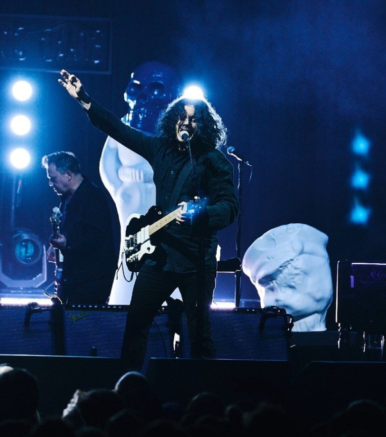 Man dressed in all black holds his right hand above his electric guitar while singing into a microphone. Another guitarist is visible in profile behind him.