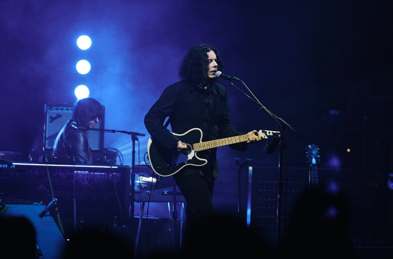Man dressed in all black strumming electric guitar stands in front of a microphone singing. 