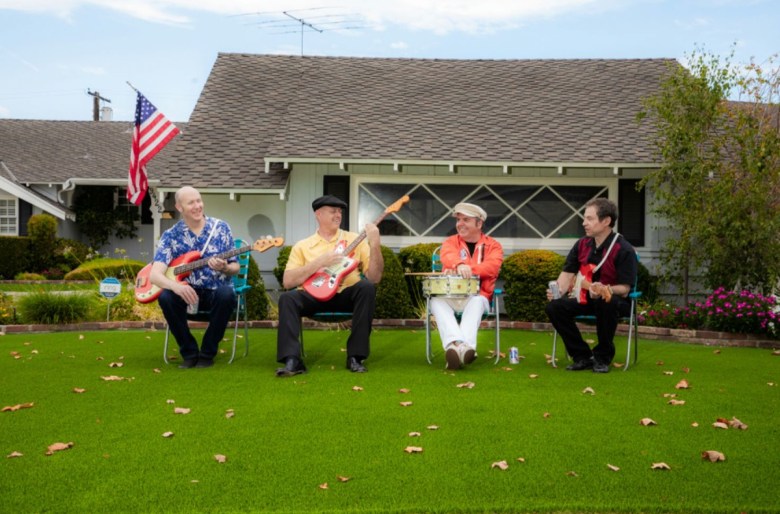Four men sit on chairs in the front yard of a home with a U.S. flag hanging over the door. The men each hold a guitar in their hands, but one man holds a single drum.