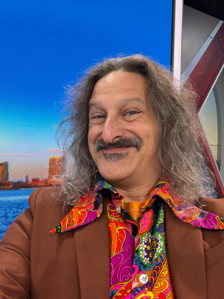 A man with long gray hair and a brief paisley dress shirt grins for the camera.