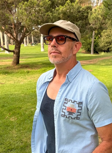 A man in a baseball cap and sunglasses looks into the distance while standing on a field of grass backed by several trees.