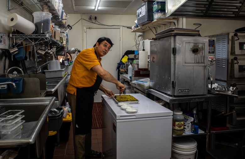 A man in a yellow shirt and black apron prepares food on a counter in a commercial kitchen, surrounded by various kitchen appliances and utensils.