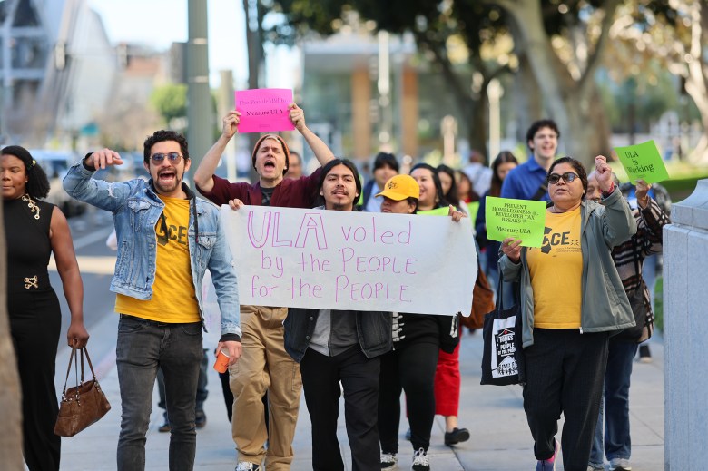 A group of people chant hold signs as they march down a sidewalk. One of the several signs reads, "ULA voted by the People for the People."