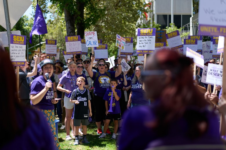 A group of people wearing purple shirts stand in a circle, chant and hold up signs that say "SEIU Local 1000" as they look and listen to a person speaking into a microphone.