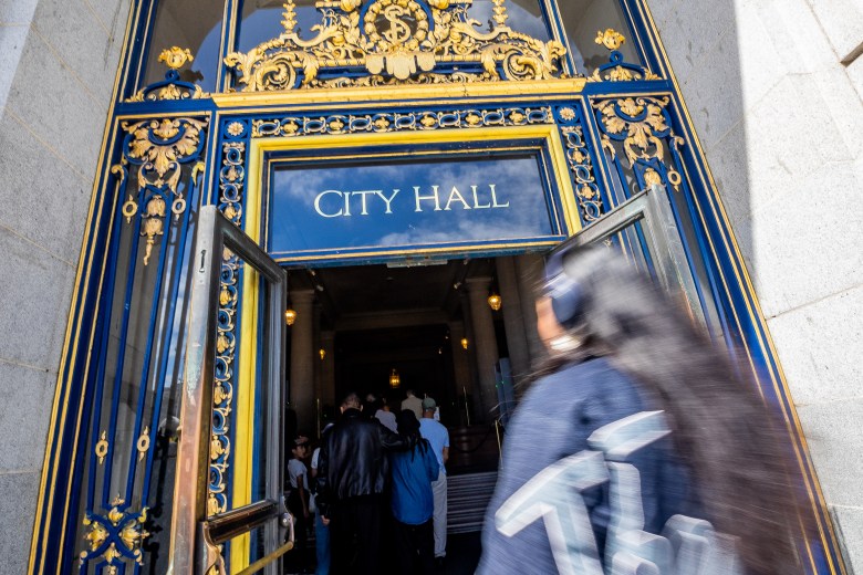 People enter a building with ornate blue and gold doors labeled “City Hall” above the entrance.