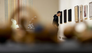 A student in a black hoodie studies a row of framed works on a gallery wall, seen past blurred objects in the foreground, with a tall multicolored hanging piece and small sculptures on a pedestal behind him.