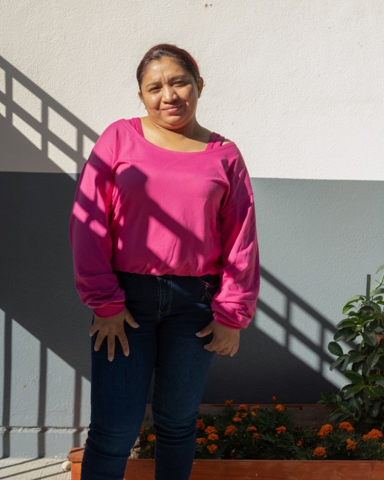 A woman in a bright pink top and dark jeans stands smiling in strong sunlight, with diagonal railing shadows across her and a flower bed behind her.