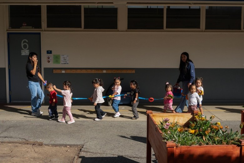Preschool children follow a blue rope in a line across a sunny schoolyard, guided by two adults; a flower planter brightens the foreground.
