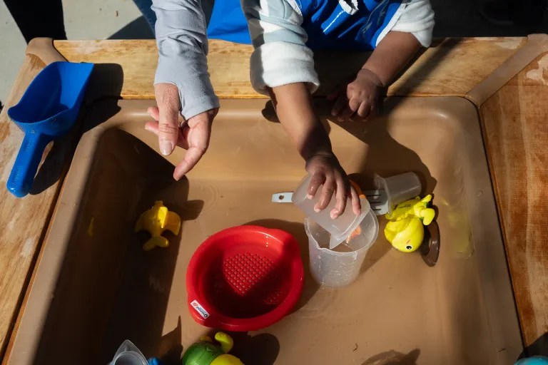 Overhead view of children exploring a water table, pouring with a clear cup among yellow duck toys, a red strainer, and blue scoop; playful learning.