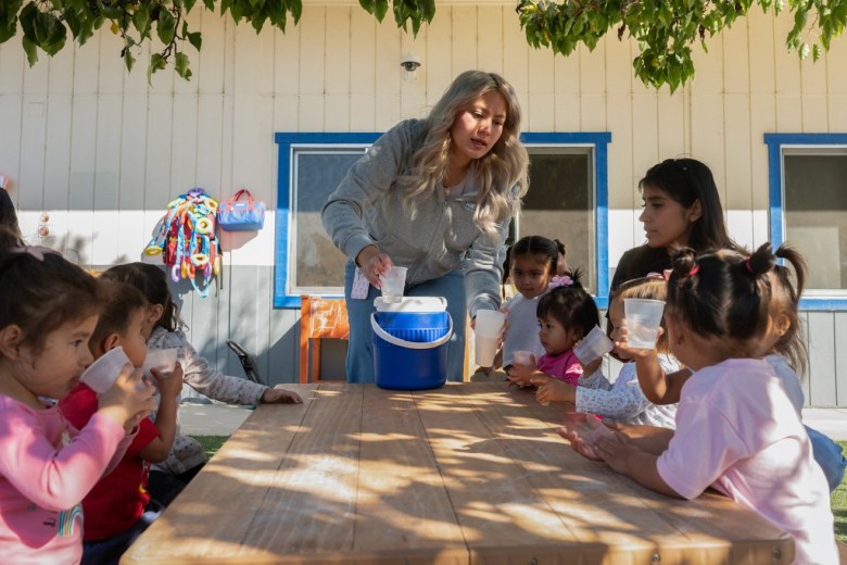 A woman pours water into a blue cooler as children gather around a wooden table, watching attentively in a sunny courtyard.