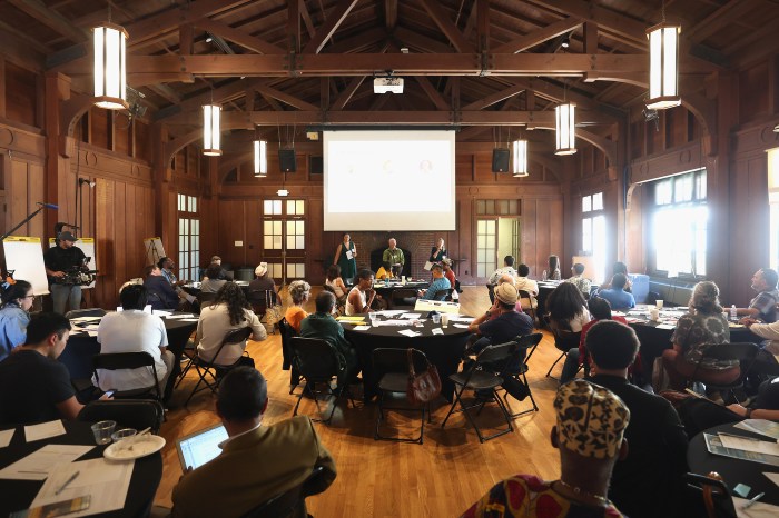 Wide view of a wood-beamed hall with hanging lanterns, where attendees sit at round tables as presenters speak at the front beside a projection screen.