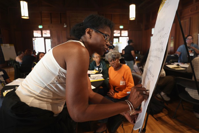 A participant leans in to write ideas on an easel pad as two women seated at her table watch, with other groups working at tables throughout the hall.