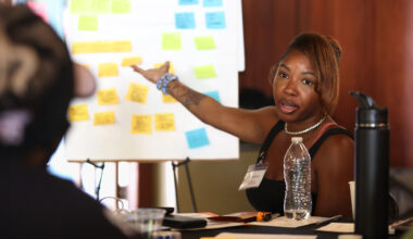 A participant gestures toward an easel pad covered in sticky notes while talking with someone across the table.