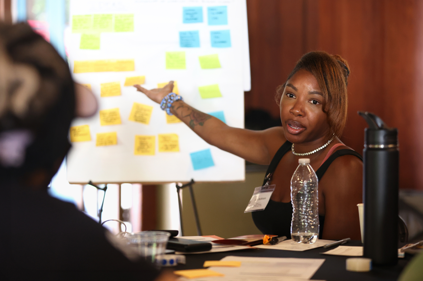 A participant gestures toward an easel pad covered in sticky notes while talking with someone across the table.