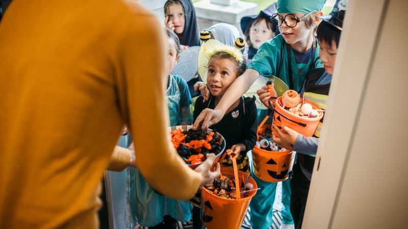 A cute multiethnic group of young elementary age kids trick or treating for the Halloween holiday in their neighborhood. They wear assorted traditional costumes, carrying Jack-o'-lantern buckets filled with candy, treats, and toys. A woman hands them candy from the front door of her home.