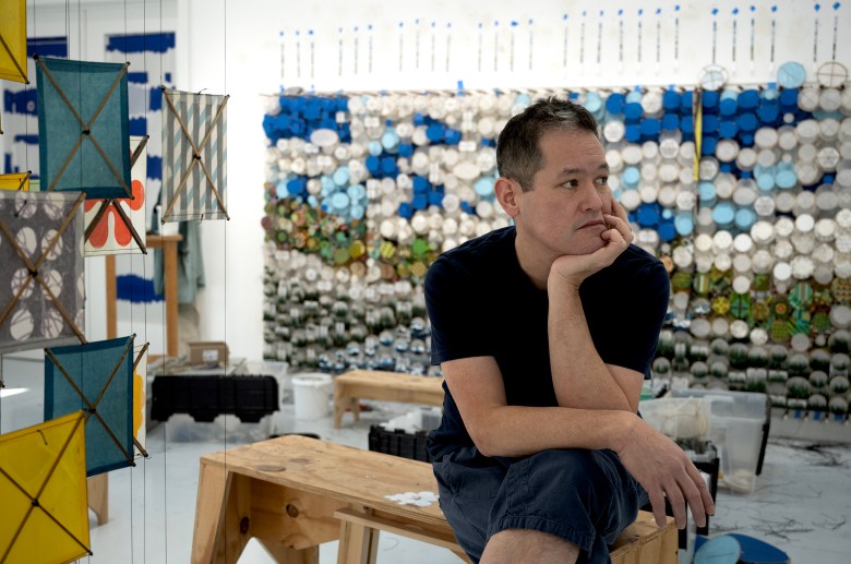 A man sits on a wooden bench in an art studio, looking thoughtful. Colorful geometric art pieces hang nearby; a blue and white wall installation fills the background.
