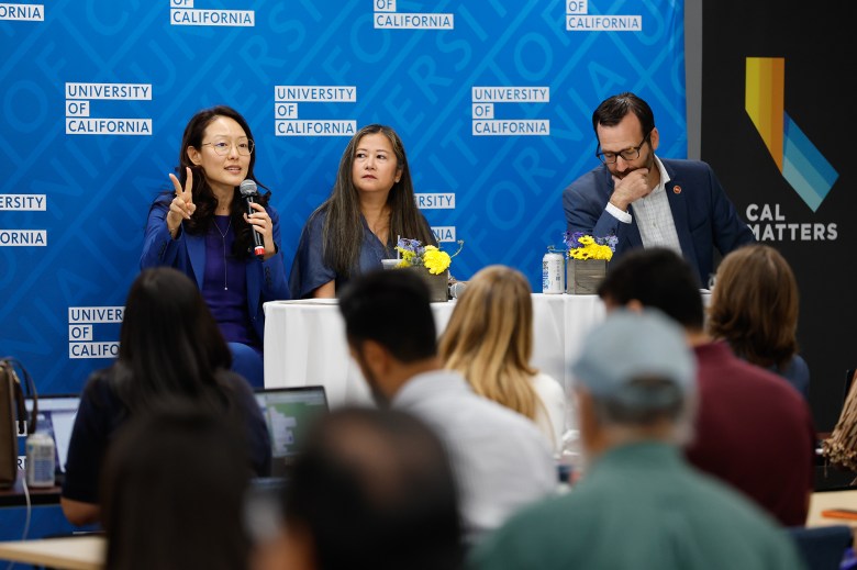 Three people sit in front of a roundtable overlooking a small room with attendees. The person on the far left gestures with their right hand as they speak into a microphone. The person in the center looks to their right and the person on the far right rest their hand on their chin as they look down. A blue step-and-repeat with the University of California logo can be seen in the background.
