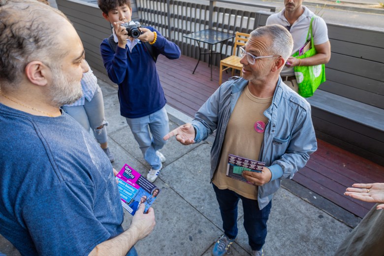 A man in a light blue jacket gestures while speaking to another man holding a political flyer, as a photographer in the background takes a picture.