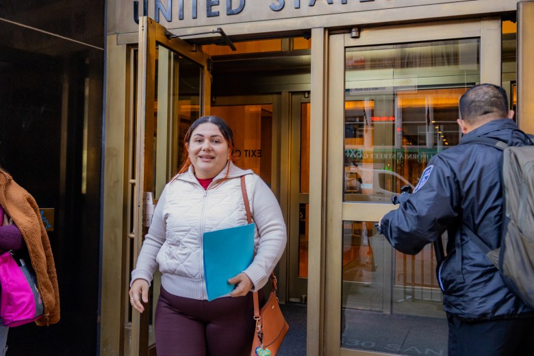 A San Francisco educator holding a blue folder exits a building with “UNITED STATES” lettering above the doorway, while a security officer stands to her right.