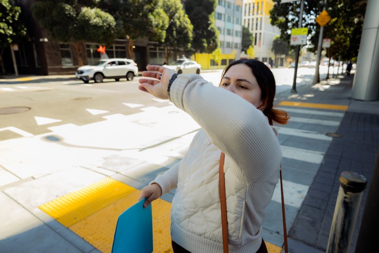 A San Francisco educator standing on a city street corner, shielding her face from the sun with her arm while holding a blue folder.