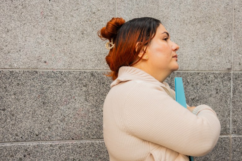 A San Francisco educator with auburn hair in a bun stands in profile against a gray stone wall, holding a blue folder under her arm.