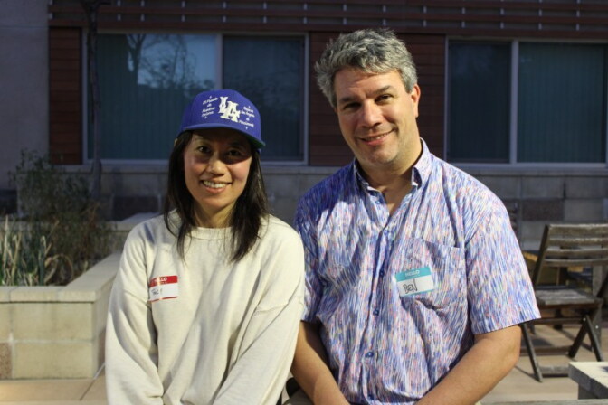 A woman with medium skin tone, wearing a cream-colored sweater and blue hat, smiles and poses for a photo with a man with light skin tone, wearing a patterned short-sleeve shirt, as they both stand outside a building.