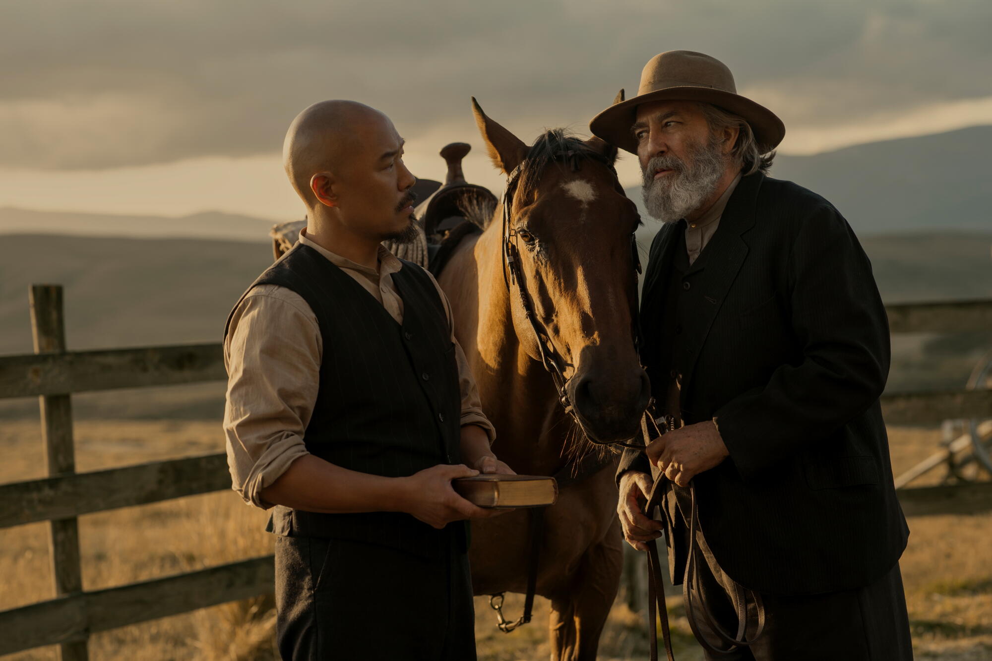 A bald man stands near the horse of a bearded man wearing a hat.