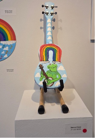 A blue ukulele painted with rainbow, cloud and cartoon frog who is playing a ukulele