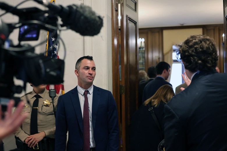 A person in a suit stands in a hallway with cameras and people around, appearing to participate in a media event or interview. An American flag and a uniformed officer are also visible.