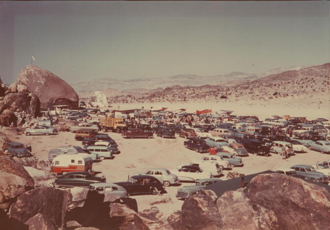 Rows of cars and trailers fill the desert beside a giant boulder with rocky hills in the background.