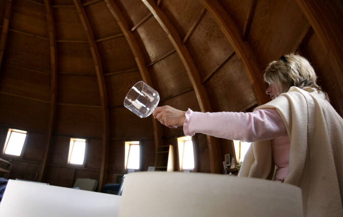 A blonde woman with her back largely to the camera hoists a glass over a quartz bowl to perform a sound bath.