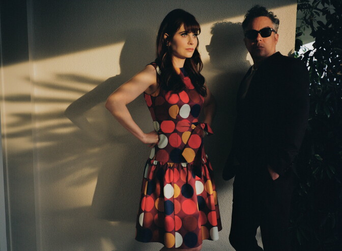 A woman in a polka-dot dress stands beside a man in sunglasses and a dark suit, sunlight casting sharp shadows on the wall.