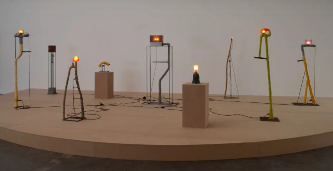 Sculptural lamps with organic, limb-like bases and glowing shades stand on a gallery platform, some on pedestals.