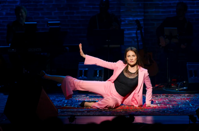 A performer in a pink suit sits on a stage rug, one leg extended, reaching an arm upward, with musicians silhouetted behind.