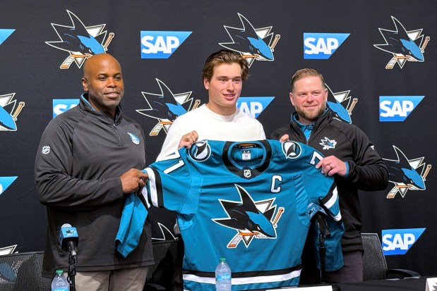 In this photo taken with a smartphone, San Jose Sharks forward Macklin Celebrini, center, is presented with the captain's jersey by general manager Mike Grier, left, and coach Ryan Warsofsky during an NHL hockey news conference in San Jose, Calif., Friday, Sept. 25, 2026. (AP Photo/Josh Dubow)